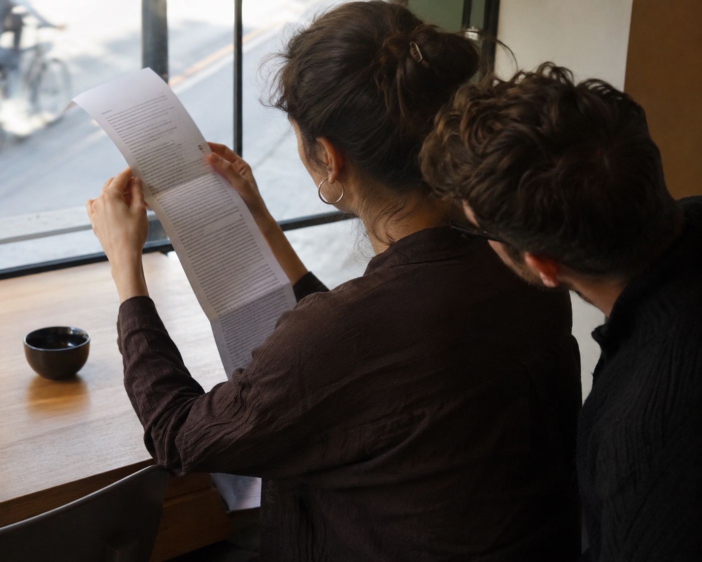 In a cafe, a woman reads a long printed letter held in both hands while a man leans in close over her shoulder, reading the same page uninvited.